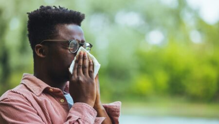 Allergic black man blowing on wipe in a park on spring season. Man with allergy or cold, blowing his nose with a tissue, looking miserable unwell very sick, isolated outside green trees background.
