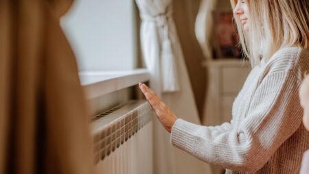 Young woman in long winter beige sweater is posing at home near the radiator