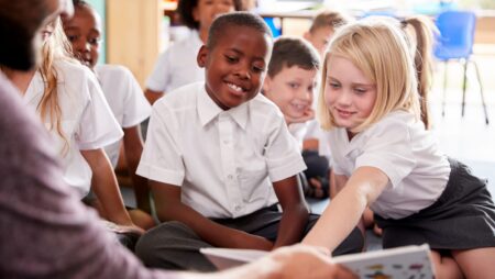 Male Teacher Reading Story To Group Of Elementary Pupils Wearing Uniform In School Classroom