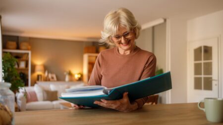 Smiling senior woman looking old photo album and reminiscing family memories while sitting at table in living room