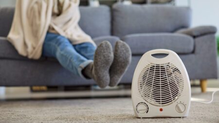 Portable fan heater heating cold home as man wrapped in blanket relaxes on sofa